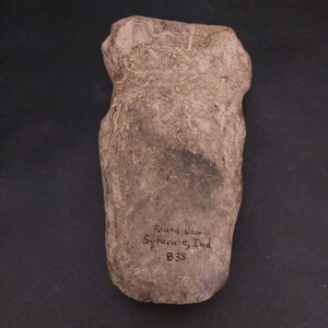 Peut inclure: Un artefact en pierre brun clair, de forme grossière et à surface texturée. Le texte "Found Near Syracuse, Ind. B38" est manuscrit sur la pierre. L'artefact est posé sur un fond sombre.