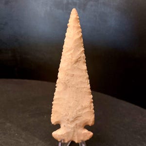 Puede incluir: Una punta de flecha de color beige con una punta triangular afilada y una base con muescas. La punta de flecha de piedra tiene una superficie texturizada. Se exhibe sobre un soporte transparente sobre un fondo oscuro. Es un artefacto histórico.