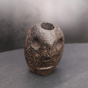 Peut inclure: Une sculpture en pierre brun foncé en forme d'œuf avec une surface texturée. La sculpture présente deux creux circulaires en forme d'yeux et une ouverture circulaire en haut. Une ligne horizontale suggère une bouche, donnant à la pierre un aspect de visage.