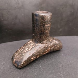 Pode incluir: Um cachimbo de pedra cinza escuro esculpido à mão com um design em forma de T. O cachimbo apresenta uma haste cilíndrica e uma base plana e texturizada. A base tem um padrão geométrico esculpido.