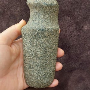Puede incluir: Una herramienta de piedra lisa de color gris, de mano, con la parte superior redondeada y la parte inferior plana. La herramienta es probablemente una piedra de moler o un martillo prehistórico.