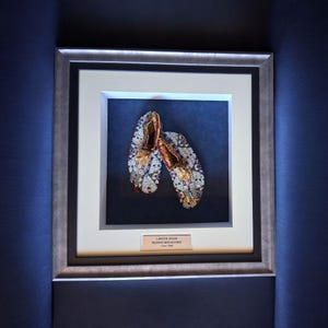 Puede incluir: Mocasines Lakota Sioux con cuentas enmarcados de 1900-1960. Los mocasines se exhiben en una caja de sombra con un marco plateado y un paspartú blanco. Los mocasines están decorados con cuentas de colores y detalles dorados. El fondo es azul oscuro.
