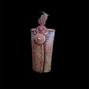 Peut inclure: Un récipient cylindrique en céramique avec une finition tachetée rouge-brun et verte. Une petite figurine ronde sculptée avec une plume attachée à la tête émerge du sommet, présentant un emblème circulaire sur la poitrine et des détails linéaires.