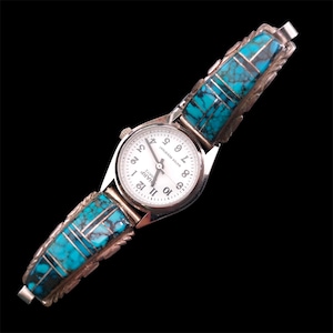 May include: Silver-toned watch with a white face and black numerals. The watch band features rectangular turquoise inlays with silver accents. The watch face reads "HARP QUARTZ" and "WATER RESISTANT".
