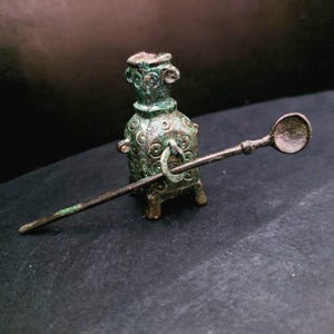 Peut inclure: Petit récipient en bronze vieilli avec un outil en forme de cuillère. Le récipient a un corps cylindrique avec des motifs circulaires décoratifs et un anneau. La cuillère est longue et fine, avec un petit bol arrondi. Le bronze a une patine verte.