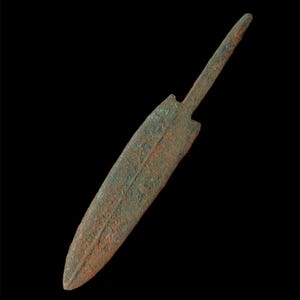 Puede incluir: Una antigua daga de bronce con una hoja en forma de hoja y un espigo rectangular. La daga tiene una apariencia desgastada y envejecida con una pátina marrón rojiza y verde, lo que sugiere su edad. El fondo es negro.