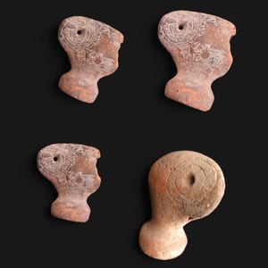 Puede incluir: Cuatro pequeñas figuras de arcilla de color terracota. Cada una tiene forma de cabeza con un agujero en la parte superior y detalles grabados. Las figuras están dispuestas sobre un fondo negro, mostrando su textura y diseño rústicos.