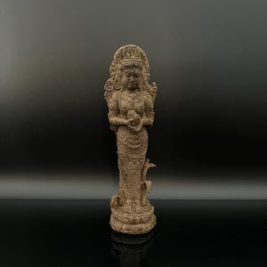 Puede incluir: Estatua detallada y desgastada de una deidad, probablemente una diosa, en posición de pie. La figura está adornada con un halo y sostiene un pequeño orbe. La estatua es de color marrón apagado y tiene detalles intrincados.