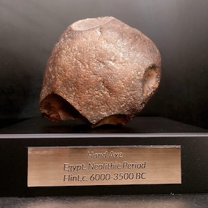 May include: A brown hand axe made of flint, dating from the Neolithic Period in Egypt, c. 6000-3500 BC. The axe has a rounded shape with a chipped edge and a hole on the side. The text "Hand Axe" and other details are on a metal plate.