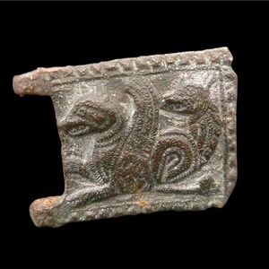 Peut inclure: Un artefact rectangulaire de couleur bronze avec une sculpture en relief détaillée de deux créatures stylisées. Le design comprend des motifs complexes et une surface texturée, sur fond noir.