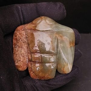 May include: A carved stone sculpture in shades of green and brown, held against a black background. The sculpture has a rounded top and a rectangular base with carved horizontal lines. The stone has a smooth, polished appearance.