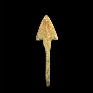 Puede incluir: Una punta de flecha de bronce con una cabeza triangular y un tallo largo y delgado. La punta de flecha tiene un aspecto desgastado y envejecido con una pátina marrón verdosa, lo que sugiere que es un artículo antiguo. El fondo es negro.