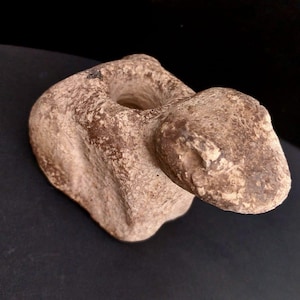 May include: A weathered, beige-colored stone sculpture with a hole in the center and a rounded, protruding shape. The sculpture has a rough, textured surface and appears to be an ancient artifact or decorative piece.
