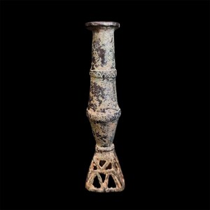 May include: A tall, cylindrical candlestick with a weathered, mottled brown and grey patina. The body features two horizontal bands with etched cross-hatch patterns, and the base is an open, triangular lattice structure with a flat top rim.