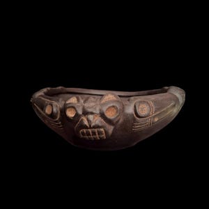 Może przedstawiać: Płytka, ciemnobrązowa ceramiczna miska z wyrzeźbioną, stylizowaną twarzą zwierzęcia na zewnętrznej stronie. Wzór zawiera okrągłe oczy i prostokątne usta z zębami w kształcie kratki, podkreślone teksturowanymi, jasnobrązowymi detalami na twarzy.