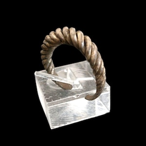 Op de afbeelding: Een zilverkleurige, gedraaide metalen armband. De armband heeft een spiraalvormig ontwerp en wordt gepresenteerd op een heldere, rechthoekige standaard tegen een zwarte achtergrond. Het ontwerp van de armband doet denken aan een touw.