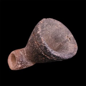 Puede incluir: Una pequeña lámpara de aceite de cerámica, hecha a mano, de color gris oscuro, con un cuenco acampanado y un pico tubular. La lámpara tiene una superficie rugosa y texturizada sobre un fondo negro.