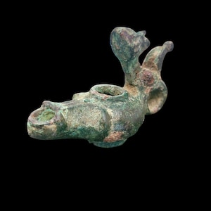 May include: An antique bronze oil lamp with a bird-like design. The lamp features a spout, a central bowl, and a handle. The bronze has a green patina, indicating age. The lamp is set against a black background.