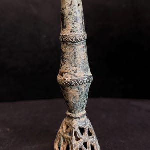Peut inclure: Un bougeoir en bronze vieilli avec une base évasée et un corps cylindrique. Le corps présente des bandes décoratives avec des motifs hachurés. Le bougeoir a une patine foncée et un petit trou près du sommet.