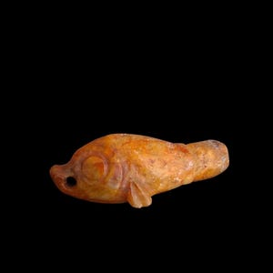 Puede incluir: Una pequeña figura de pez tallada en piedra en tonos ámbar, naranja y marrón. El objeto presenta un cuerpo redondeado, una cuenca ocular definida, una pequeña aleta en la parte inferior y un orificio circular cerca de la boca para colgar.