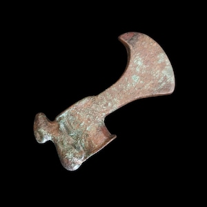 May include: A weathered, bronze-coloured axe head with a curved blade and a handle. The axe head has a rounded poll and a slightly curved blade. The metal has a mottled appearance, suggesting age and use. The background is black.