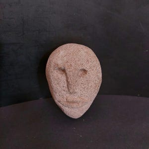 May include: A small, oval-shaped sculpture of a face. The sculpture is a light brown color with a textured surface. The eyes, nose, and mouth are subtly carved into the stone. The background is a dark, solid color.