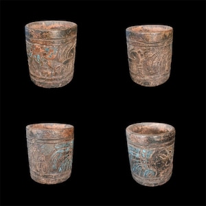May include: Four weathered, cylindrical vessels with carved relief designs. The vessels are primarily brown with hints of teal and rust-colored accents. The relief carvings appear to depict stylized figures and patterns. The background is black.