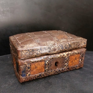 Puede incluir: Un pequeño cofre metálico antiguo de forma rectangular. El cofre es principalmente marrón con detalles dorados y óxido. La tapa y los lados están decorados con tiras y remaches metálicos, y un ojo de cerradura es visible en la parte delantera.