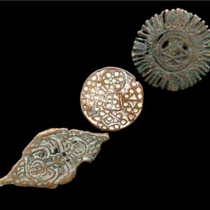 Könnte beinhalten: Drei antike Bronze-Artefakte auf schwarzem Hintergrund. Die Artefakte umfassen ein sonnenförmiges Medaillon, ein kreisförmiges Stück mit floralem Muster und ein blattförmiges Stück mit Spiralmuster. Die Bronze hat ein verwittertes, gealtertes Aussehen.