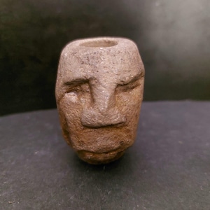May include: A small, weathered stone sculpture of a human face. The face has closed eyes, a simple nose, and a slightly open mouth. The sculpture is a light brown color and has a cylindrical shape with a hole at the top.