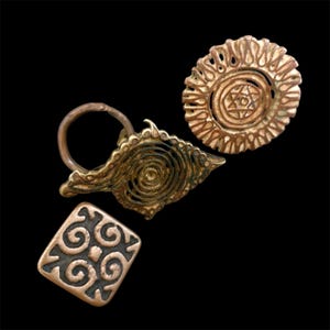 Könnte beinhalten: Drei antike Schmuckstücke aus bronzefarbenem Metall auf schwarzem Hintergrund. Die Stücke umfassen einen runden Anhänger mit einem Stern, einen spiralförmigen Anhänger mit einem Ring und einen quadratischen Anhänger mit einem Wirbelmuster. Der Schmuck hat ein gealtertes Aussehen.