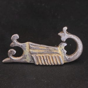 Könnte beinhalten: Eine silberfarbene Metallbrosche mit einem komplizierten Design. Die Brosche ist in Form eines stilisierten Vogels mit einem langen, geschwungenen Schwanz geformt. Der Körper des Vogels ist mit einer Reihe von parallelen Linien verziert.