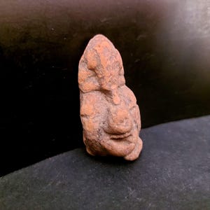 Puede incluir: Pequeña escultura de color marrón rojizo de un rostro humano. La cara tiene rasgos definidos, incluyendo nariz, boca y ojos. La escultura parece estar hecha de un material rugoso y texturizado, posiblemente arcilla o piedra. El fondo es oscuro.