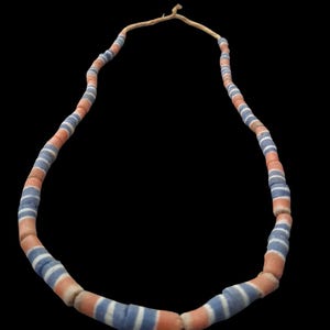 Peut inclure: Un collier composé de perles cylindriques alternant le bleu, le blanc et l'orange. Les perles sont enfilées sur un cordon fin et brun clair. Le collier est présenté sur un fond noir.