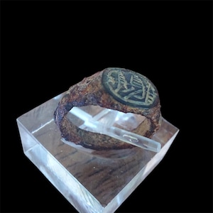 Puede incluir: Un anillo antiguo y desgastado con un emblema ovalado oscuro. El anillo es de color marrón óxido y parece estar hecho de metal. El emblema presenta un diseño intrincado. El anillo se exhibe sobre un soporte de acrílico transparente.