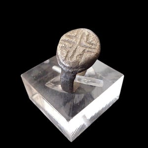 May include: A silver ring with a circular face, displaying a cross pattern. The ring is presented on a clear, square stand against a black backdrop. The ring's band is dark, contrasting with the lighter face.