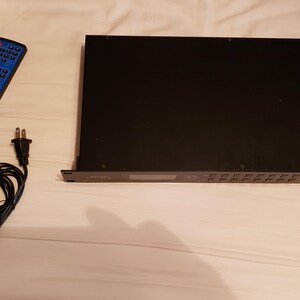 Puede incluir: Un dispositivo electr&oacute;nico negro con m&uacute;ltiples botones y una pantalla, acompa&ntilde;ado de un mando a distancia azul, un adaptador de corriente y un cable de alimentaci&oacute;n. Los art&iacute;culos est&aacute;n dispuestos sobre una superficie blanca.