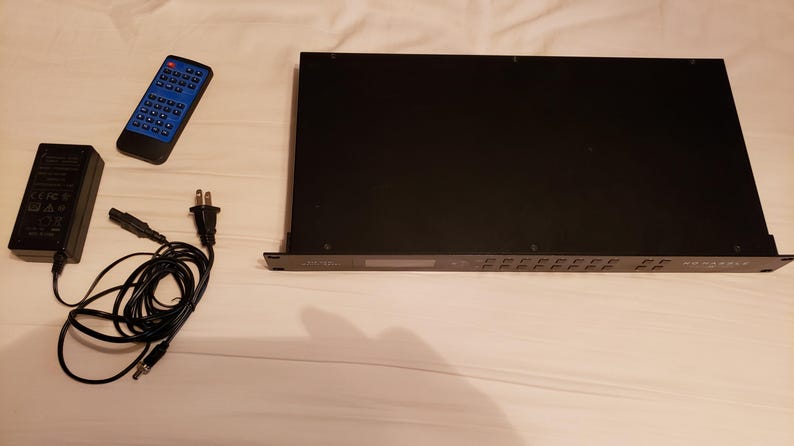 Puede incluir: Un dispositivo electr&oacute;nico negro con m&uacute;ltiples botones y una pantalla, acompa&ntilde;ado de un mando a distancia azul, un adaptador de corriente y un cable de alimentaci&oacute;n. Los art&iacute;culos est&aacute;n dispuestos sobre una superficie blanca.