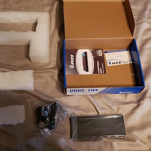 Puede incluir: Una caja de cartón abierta que contiene un dispositivo electrónico negro, materiales de embalaje y un adaptador de corriente. La caja tiene una franja azul y el texto "UHDS-104". El dispositivo está empaquetado con inserciones de espuma.