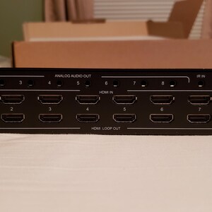 Puede incluir: Dispositivo electr&oacute;nico negro con m&uacute;ltiples puertos HDMI etiquetados 1-8, HDMI IN, HDMI LOOP OUT y ANALOG AUDIO OUT. Tiene un puerto de actualizaci&oacute;n y un puerto serie. El dispositivo est&aacute; sobre una superficie blanca.