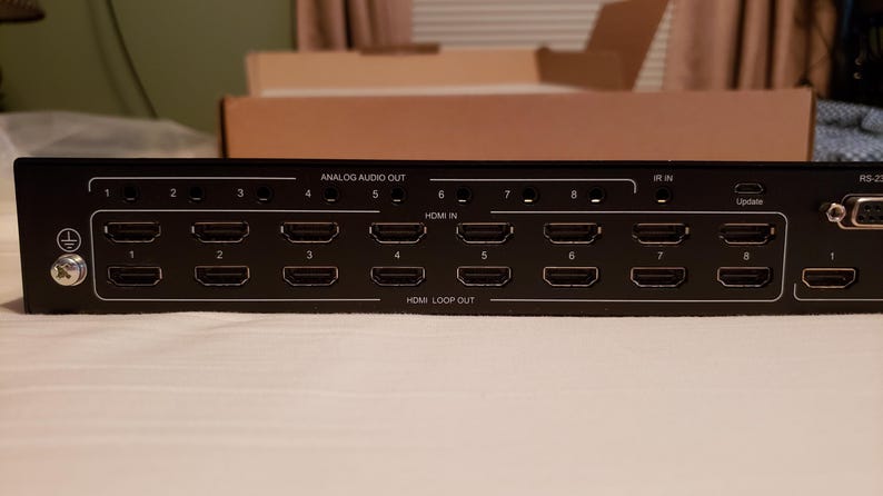 Puede incluir: Dispositivo electr&oacute;nico negro con m&uacute;ltiples puertos HDMI etiquetados 1-8, HDMI IN, HDMI LOOP OUT y ANALOG AUDIO OUT. Tiene un puerto de actualizaci&oacute;n y un puerto serie. El dispositivo est&aacute; sobre una superficie blanca.