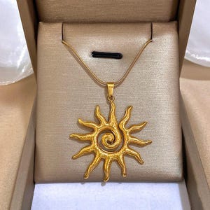 Puede incluir: Un collar de oro con un colgante de sol. El colgante es un sol estilizado con una espiral en el centro.