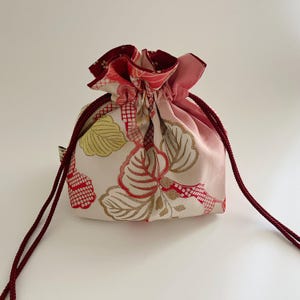 Peut inclure: Un petit sac à cordon avec un motif floral rouge, doré et jaune sur fond crème. L'intérieur est rose et il possède un cordon de serrage rouge. Le sac est probablement utilisé pour transporter de petits objets.