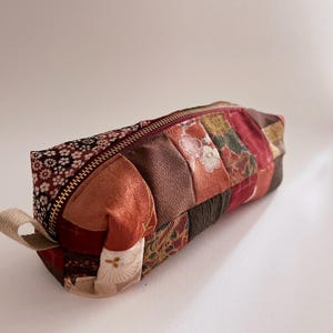 Könnte beinhalten: Ein rechteckiges Federmäppchen mit Patchwork-Design. Das Etui weist Stoffe in Rot-, Braun- und Goldtönen mit floralen und geometrischen Mustern auf. Es hat einen bordeauxfarbenen Reißverschluss und eine kleine Stoffschlaufe an einem Ende.