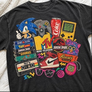 May include: A black t-shirt featuring a graphic print of 90s pop culture icons including Sonic the Hedgehog, a Nintendo 64 controller, a Coke can, a Game Boy, an MTV logo, a cassette tape labeled 90's, a sneaker, sunglasses, and a Tamagotchi.