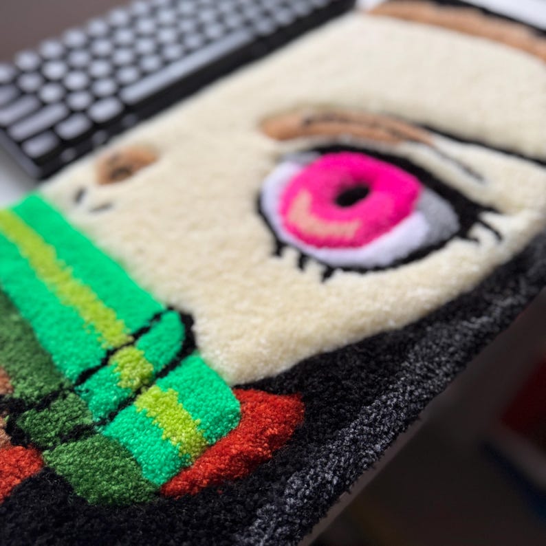 A close-up of a handmade rug featuring a nezuko’s face. The rug has a cream-colored background with a large pink eye, brown eyebrow, and green and red accents. The rug is made of a soft, textured material.