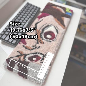 May include: A rectangular rug with a cartoon face design, measuring 19.7" x 7.5". The rug features a light pink background with brown, red, and white details. A black keyboard is visible in the background.