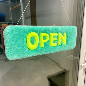 Op de afbeelding: Een turquoise rechthoekig bord met het woord "OPEN" in felgeel. Het bord hangt aan een wit touwtje, wat aangeeft dat de winkel open is.