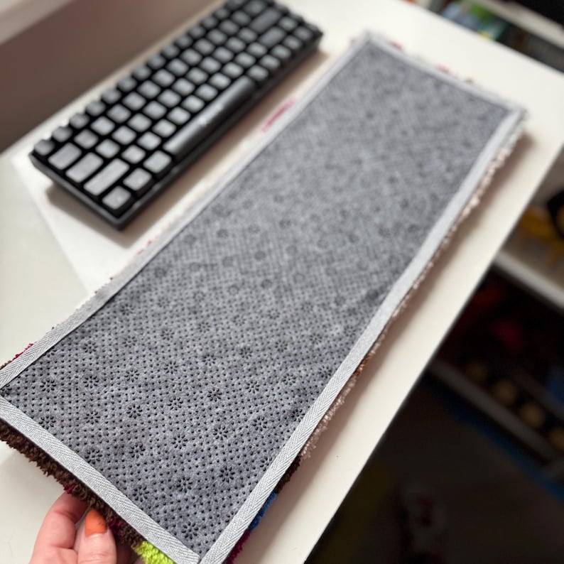 May include: A rectangular rug with a gray, non-slip backing and a colorful border. A black keyboard is in the background. The rug's border features a mix of colors, including brown, green, and blue.