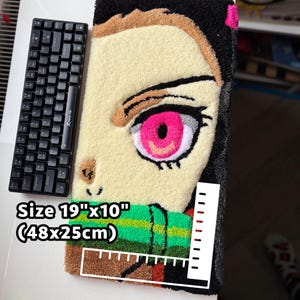 May include: A handmade rug featuring a character's face with a large pink eye, measuring 19" x 10". The rug has a black border and a green and brown scarf. A black keyboard is placed next to the rug.